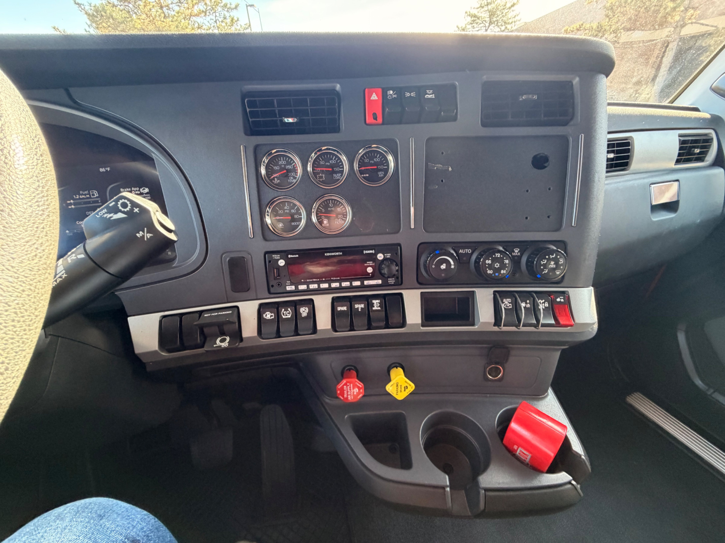 Kenworth T680 dashboard showing multiple gauges, switches, and controls. Center stack with round gauges, hazard button, climate controls, radio, and red ignition switch. Yellow and red control levers at bottom.
