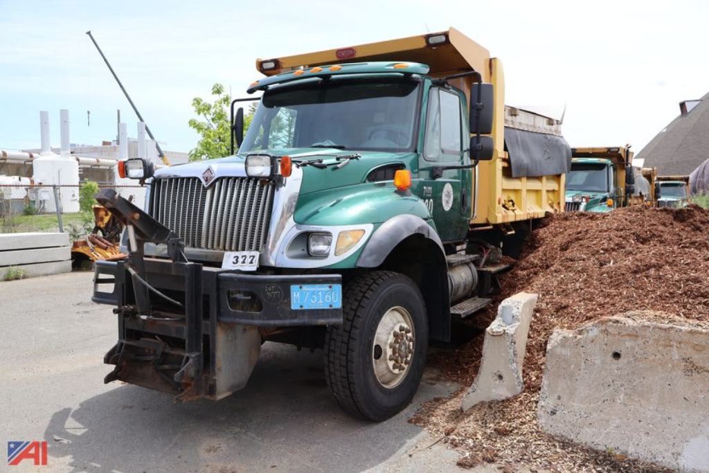 International WorkStar 7400 dump truck in a yard, green cab, yellow dump bed, front hitch, and pile of dirt beside it.
