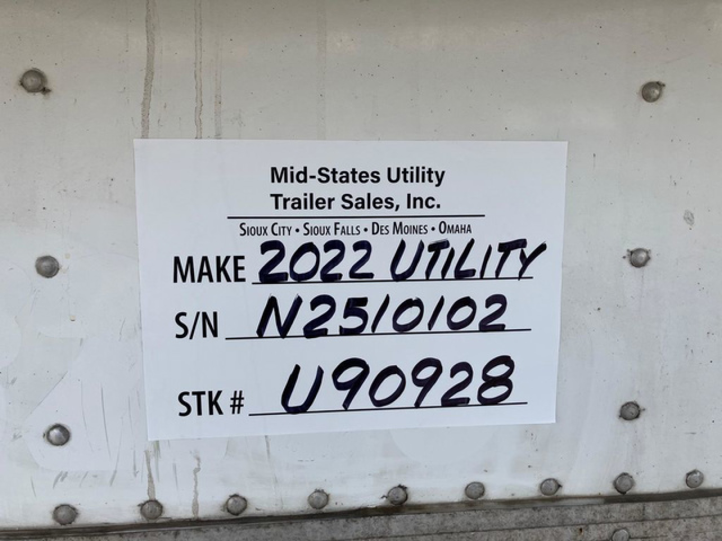 Trailer side with white label reading Mid-States Utility Trailer Sales, Inc. Make 2022 Utility, S/N N2510102, STK # U90928. VIN printed elsewhere; surroundings show riveted metal of refrigerated trailer.