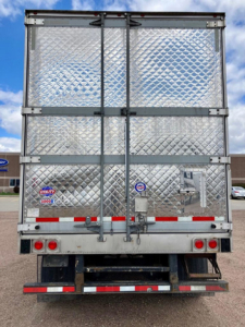 Rear view of a 2022 utility reefer trailer with diamond plate aluminum doors, hinges, and latches. Stocked with reflective red and white tape, taillights, and undercarriage visible.