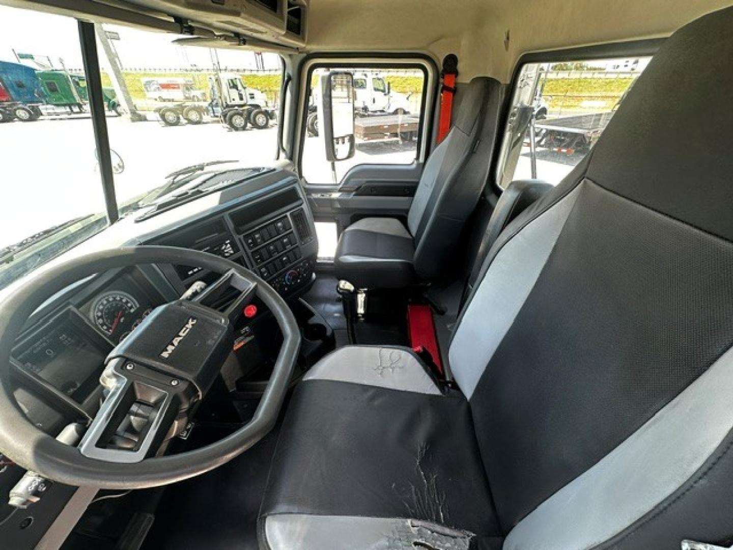 Mack PI64T interior showing steering wheel, dashboard, driver seat, and passenger seat in cab; visible windows and exterior trucks in lot.