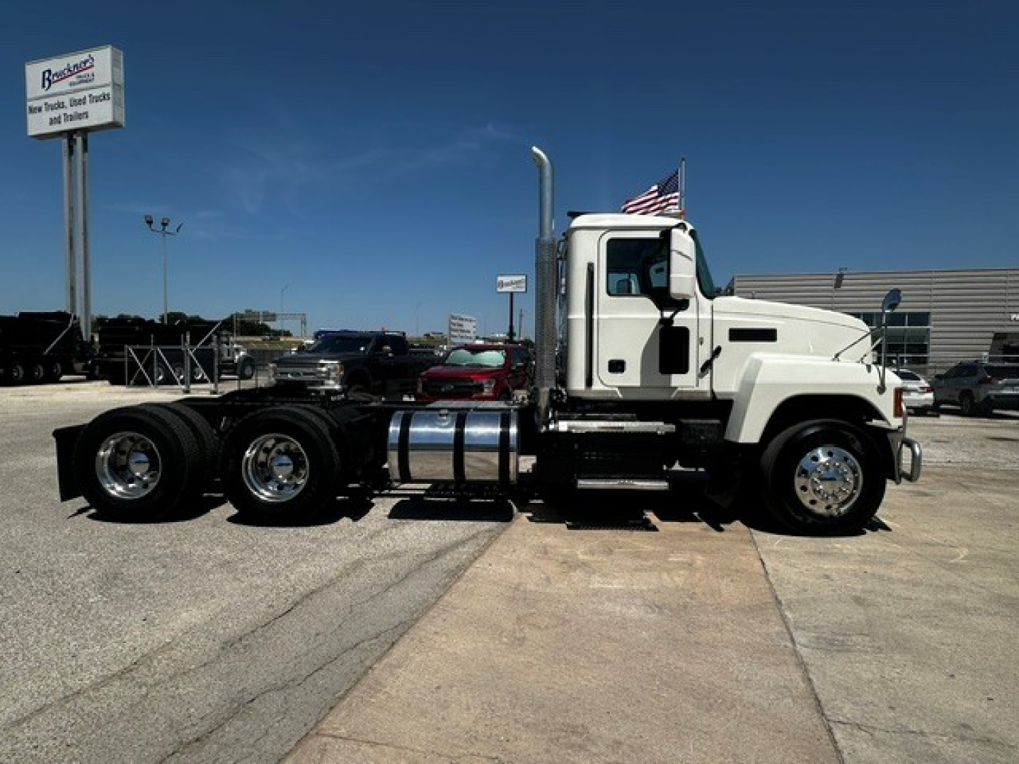 Truck side view showing a white 2020 Mack PI64T conventional daycab with three rear axles and chrome fuel tanks. Visible text includes a dealership sign in the background and an American flag on the cab.