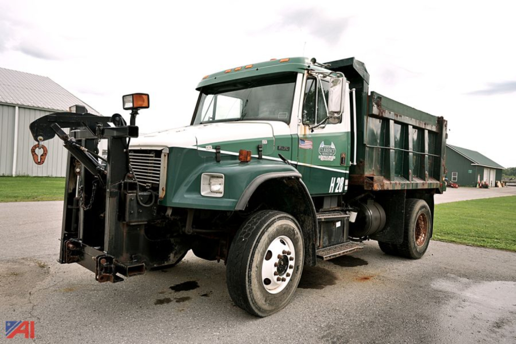 Freightliner FL70 dump truck with plow attachment in a yard, green cab with white top, H28 badge on door, dump bed in rear