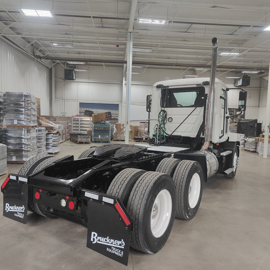 Side view of a 2020 Mack AN64T conventional daycab tractor inside a showroom with dual rear axles and tall exhaust stack.