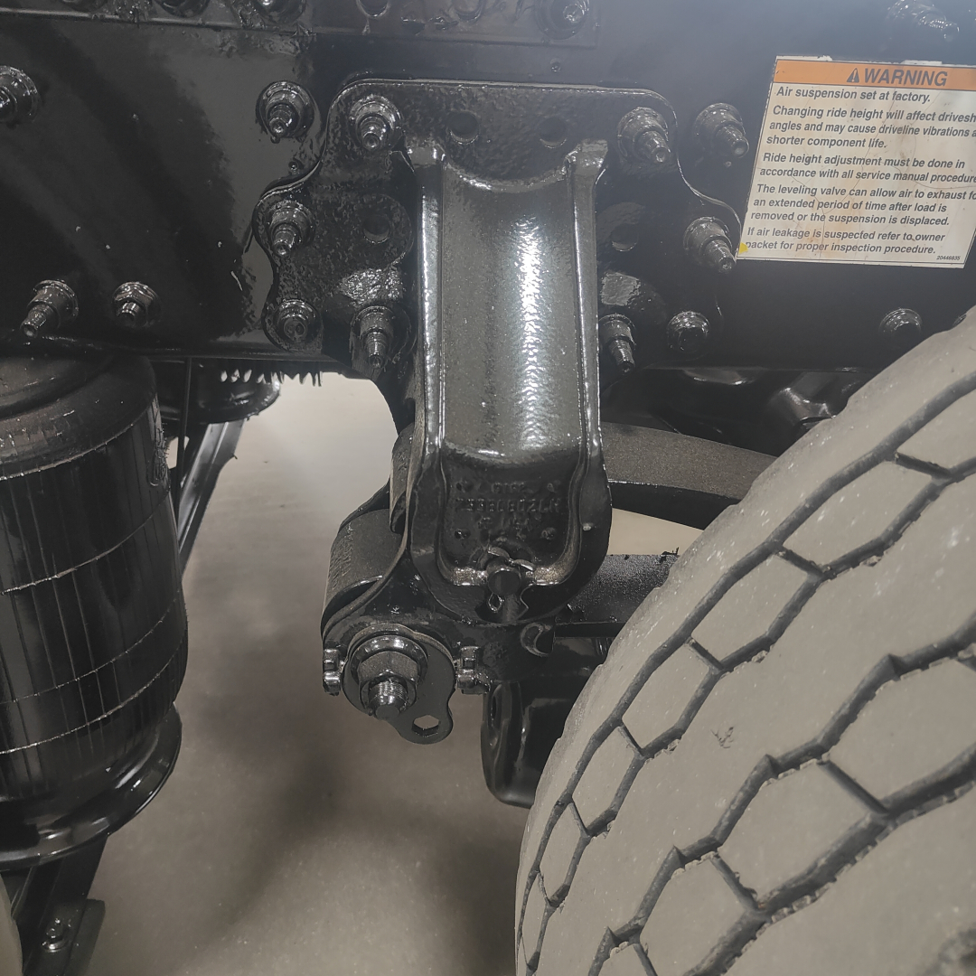 Mack AN64T chassis view showing suspension mount and front tire; focus on front axle, coil/leaf suspension bracket, and bolts; warning label visible on frame.