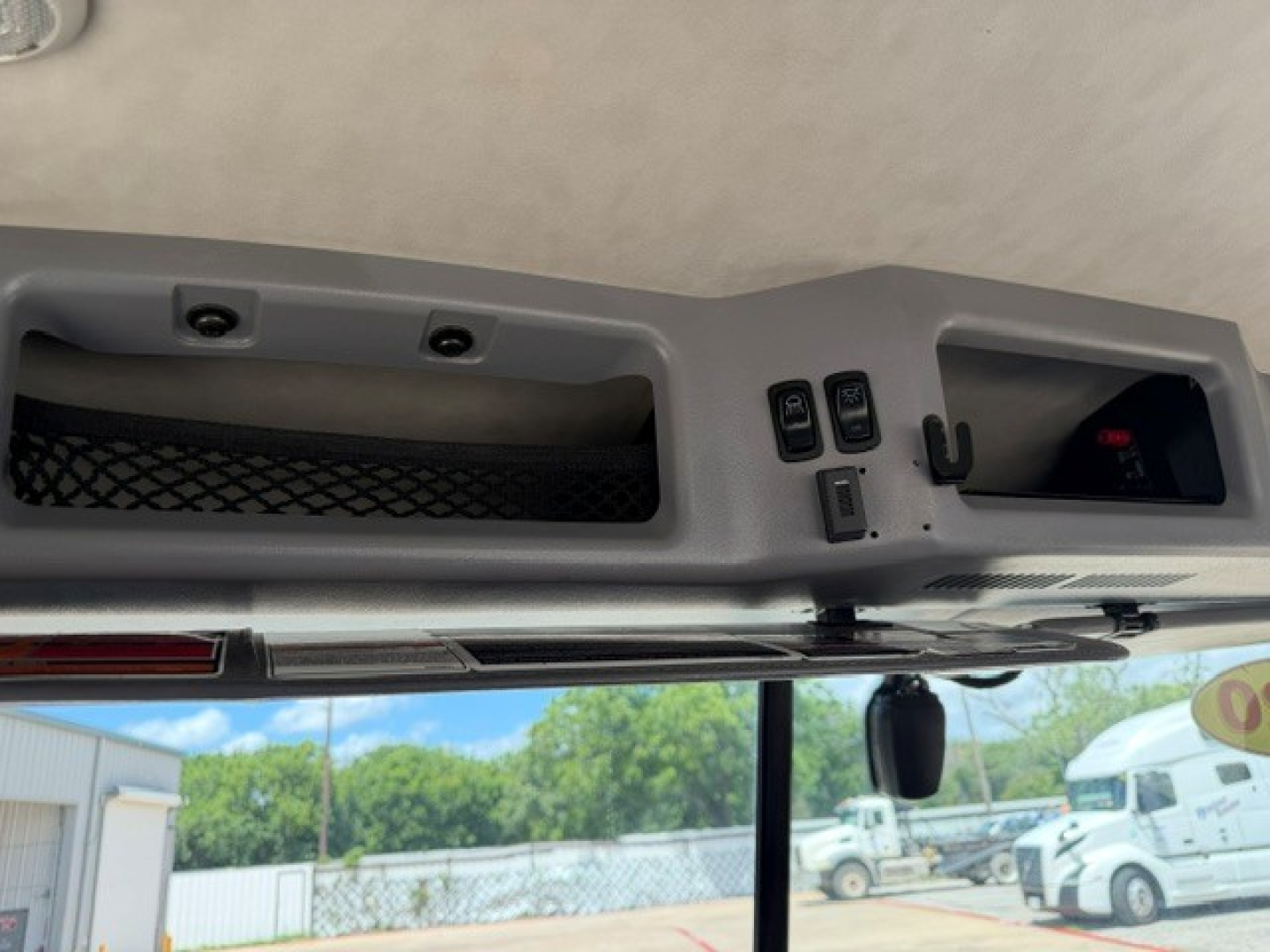 Cockpit view of a 2020 Mack AN64T interior showing overhead console with switches, small display, and mirror; windshield with exterior trucks in background.