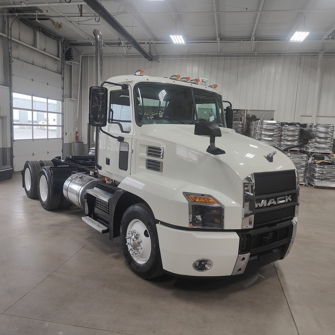 Truck engine bay side view showing Mack AN64T cab and chassis in a showroom, with white cab, black grille, and front bumper; wheel rims visible.