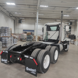 Side view of a 2020 Mack AN64T conventional daycab tractor inside a showroom with dual rear axles and tall exhaust stack.