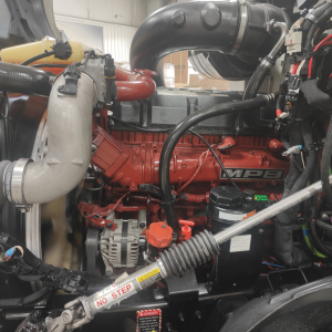 Engine bay of a Mack AN64T truck showing red diesel engine, black hoses, orange oil cap, alternator, turbo components, and label reading ‘NO STEP’.