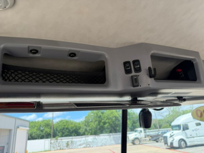 Cockpit view of a 2020 Mack AN64T interior showing overhead console with switches, small display, and mirror; windshield with exterior trucks in background.