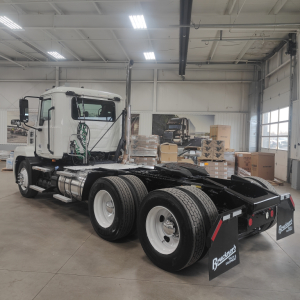 Tractor truck chassis and cab of a 2020 Mack AN64T with dual rear axles in a dealership service bay. Visible fuel tanks, cab doors, and rear wheel assembly; shop in background with boxes.