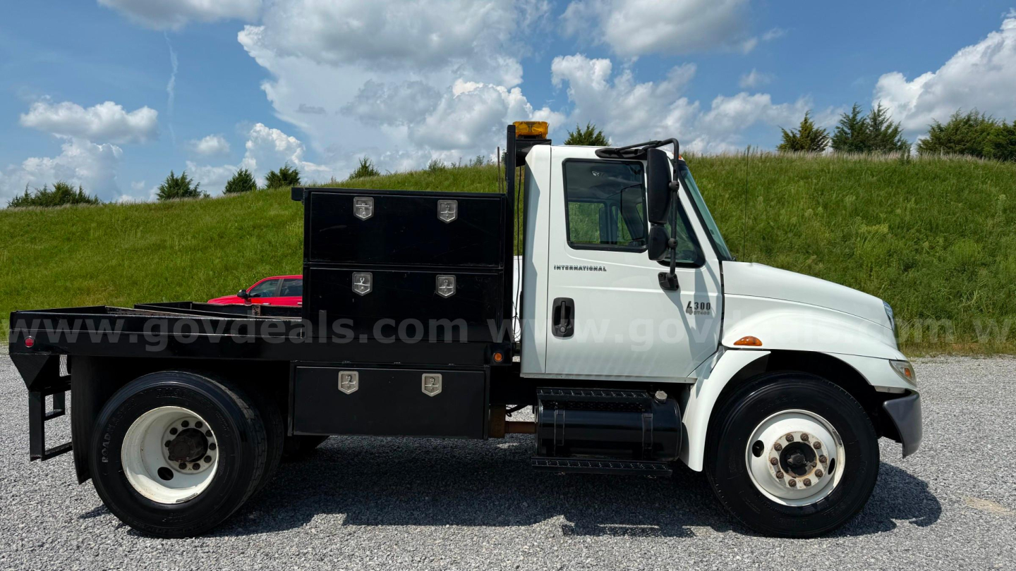 International 4300 flatbed truck side view showing tool boxes and cab, used 2007 model with white cab and black flatbed accessories.