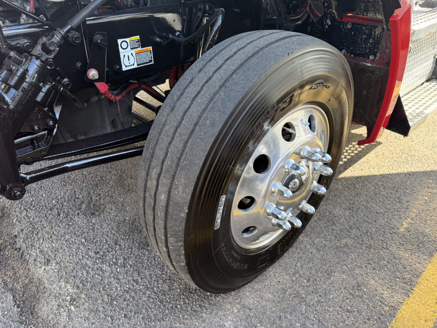 Kenworth T880 front wheel and axle assembly with dual rear axles visible, glossy chrome lug nuts, tire, and part of red cab; engine bay components and warning labels in background.