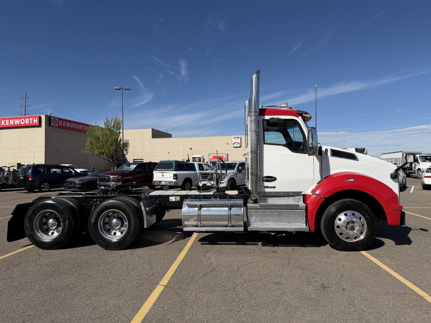 Kenworth T880 truck in a parking lot side view showing white cab with red front fenders, chassis, and dual rear axles. Clear blue sky and dealership building in background with Kenworth signage.