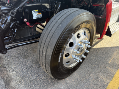 Kenworth T880 front wheel and axle assembly with dual rear axles visible, glossy chrome lug nuts, tire, and part of red cab; engine bay components and warning labels in background.