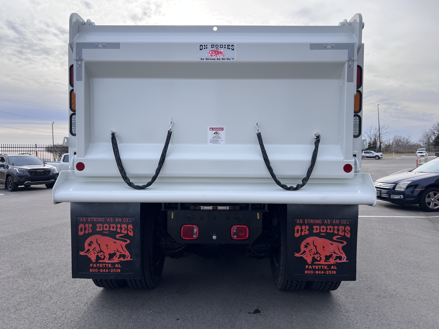 Rear view of a white Mack GR64F dump truck/ trailer showing the dump bed with two black straps, taillights, mud flaps with red graphics, and a license-free rear bumper area.