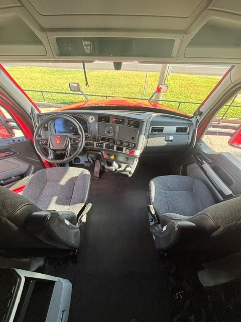 Cab interior of a Kenworth truck with steering wheel, dashboard, and two front seats. Visible controls, gauges, and cup holders, sun visors, overhead console.