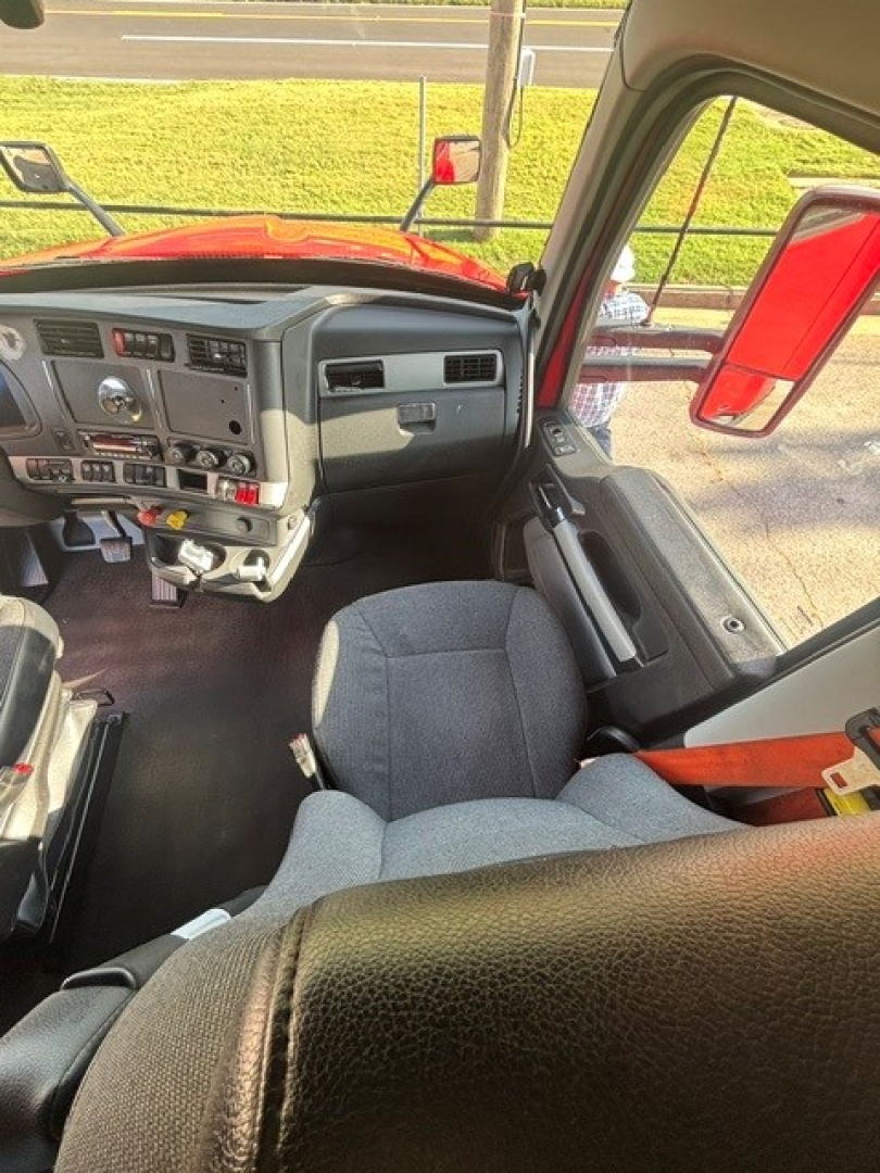 Cab interior of a 2024 Kenworth T680 showing dashboard and driver seat with steering wheel, controls, and orange seat belt in view.