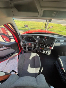Cab interior of a 2024 Kenworth T680 showing steering wheel, dashboard, and driver seat from passenger side, with red exterior visible through open door.