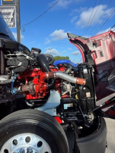 Kenworth T680 engine bay with red engine, blue hoses, and exposed front wheel; visible hood open showing engine components and surrounding chassis.