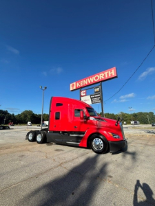 Kenworth T680 semi-truck in red paint, side profile view with sleeper cab, emblem visible, dealership sign in background.
