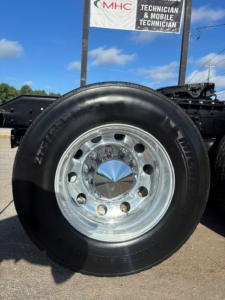 Truck wheel with polished chrome rim and black tire; portion of chassis and dealership sign in background. Visible text includes on tire side: 'M/V' and other faint markings; sign overhead shows 'MHC' and 'TECHNICIAN & MOBILE TECHNICIAN'.