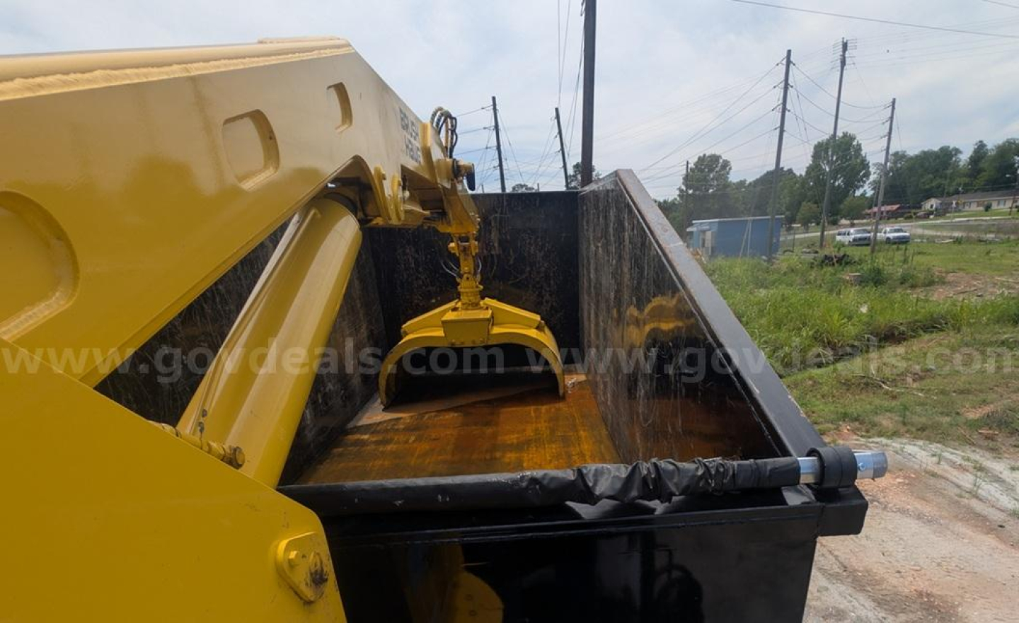 Grapple truck bed with yellow loader arm and black dump bed showing attachment and hydraulic cylinder, urban background.