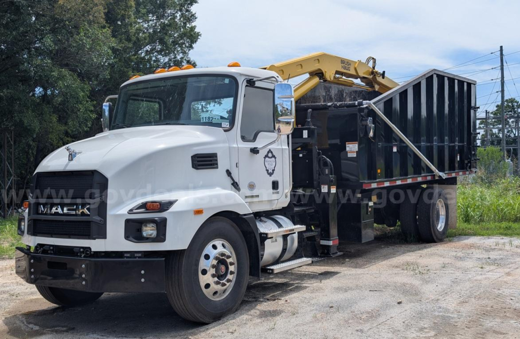 Mack MD grapple truck with a yellow hydraulic arm and black dumping bed, white cab, front grille visible.