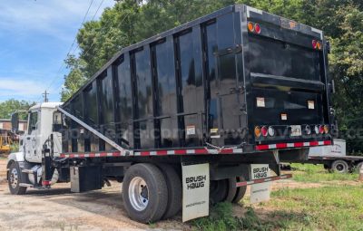 Truck rear view showing a black dump bed on a Mack MD grapple truck, dual rear wheels, mud flaps with Brush Haul branding, and safety rails along the bed.