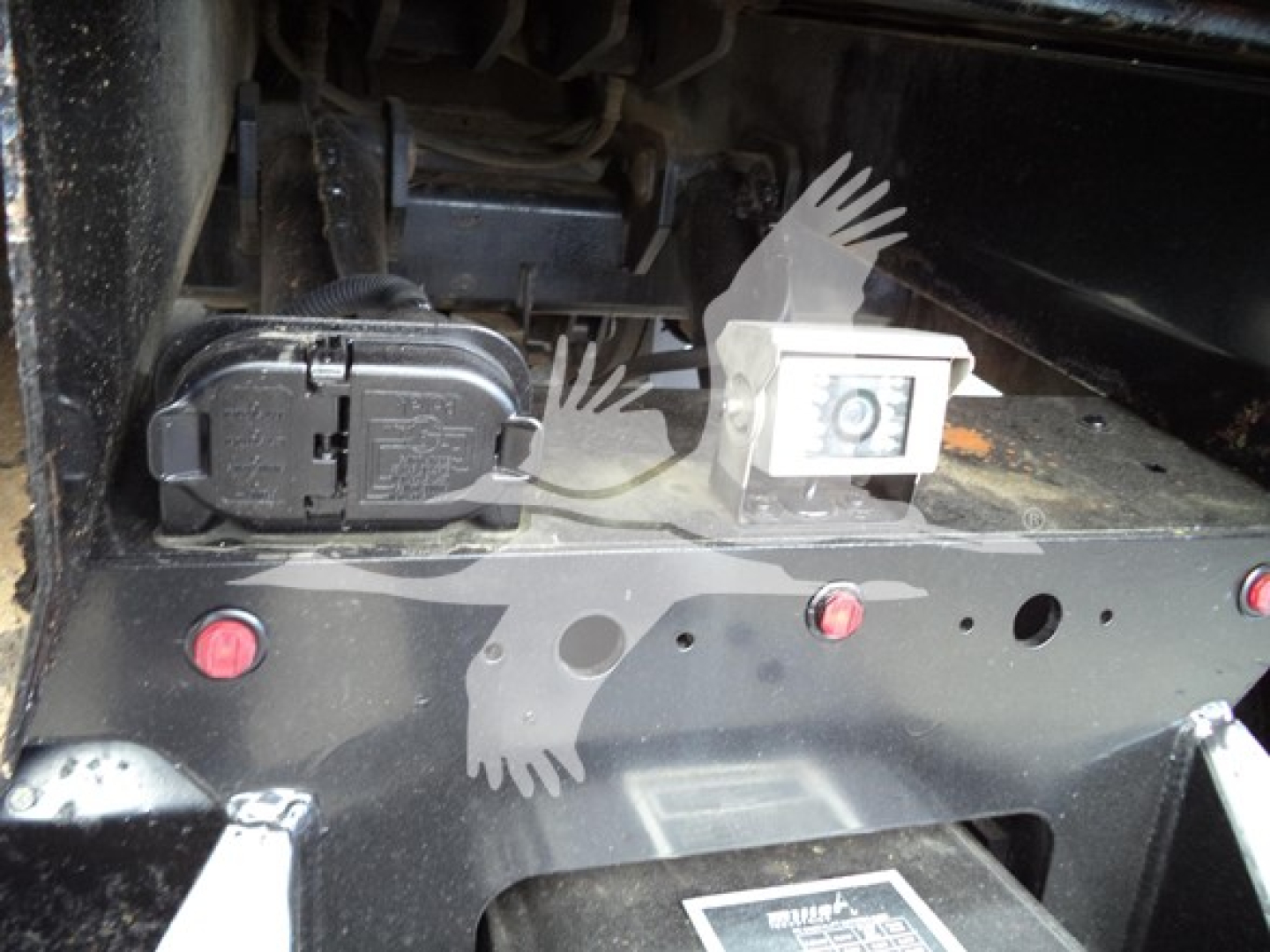 Peterbilt truck undercarriage showing electrical plug and mounted box with a white protective cap; nearby is a decorative hand stencil and fasteners.