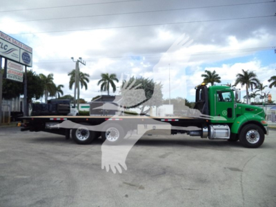 Peterbilt 348 rollback tow truck, green cab, long flatbed trailer in lot with palm trees in background. No visible text on the truck itself.