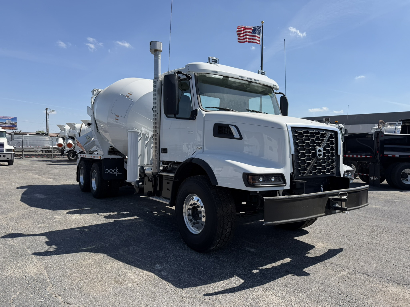 Volvo VHD64B300 mixer truck in the lot, white cab, front grille, concrete mixer drum attached, VIN 4V5KC9DH9TN706198 visible, flags in background.