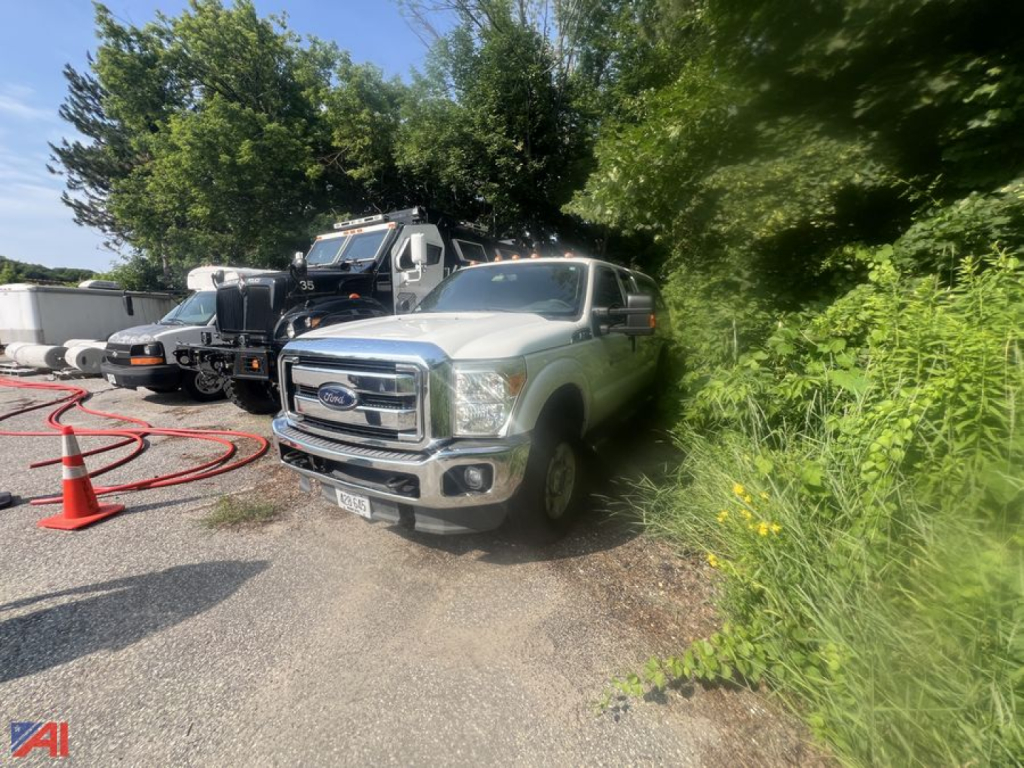Ford F-250 Super Duty pickup truck in a lot, extended cab with cap, front view, greenery on right, other vehicles in background.