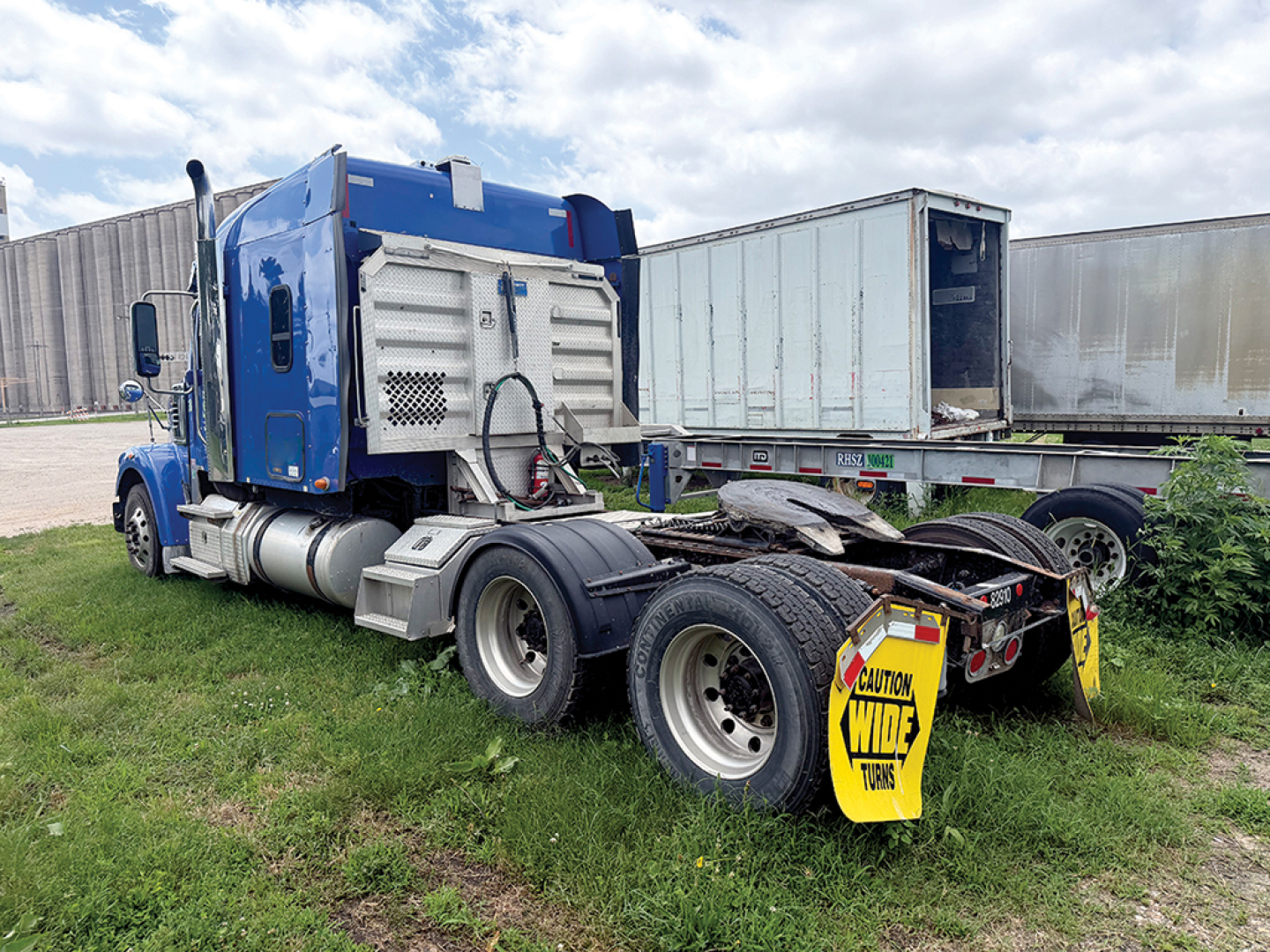 Truck cab and chassis of a blue 2018 Freightliner Coronado SD122, rear view shows fifth wheel and dual rear axles with yellow caution sign on mudflap.