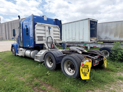 Truck cab and chassis of a blue 2018 Freightliner Coronado SD122, rear view shows fifth wheel and dual rear axles with yellow caution sign on mudflap.