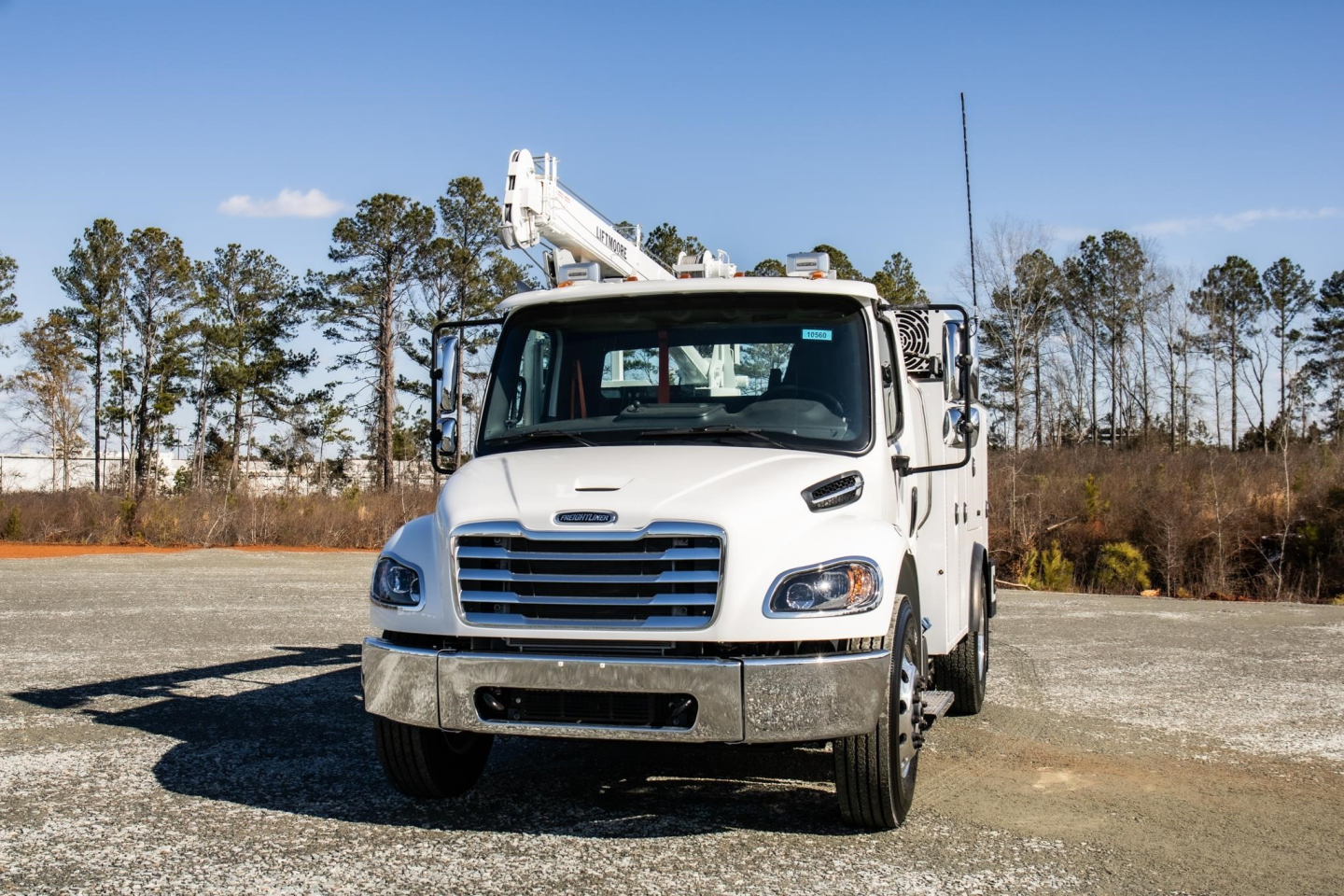 White Freightliner M2 106 Plus service truck with crane, front view, outdoor lot with trees in background.