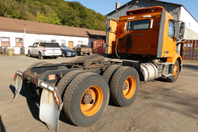 Volvo VNR64T300 conventional daycab tractor rear view showing three axles, orange cab, and black chassis with faded fuel tanks.