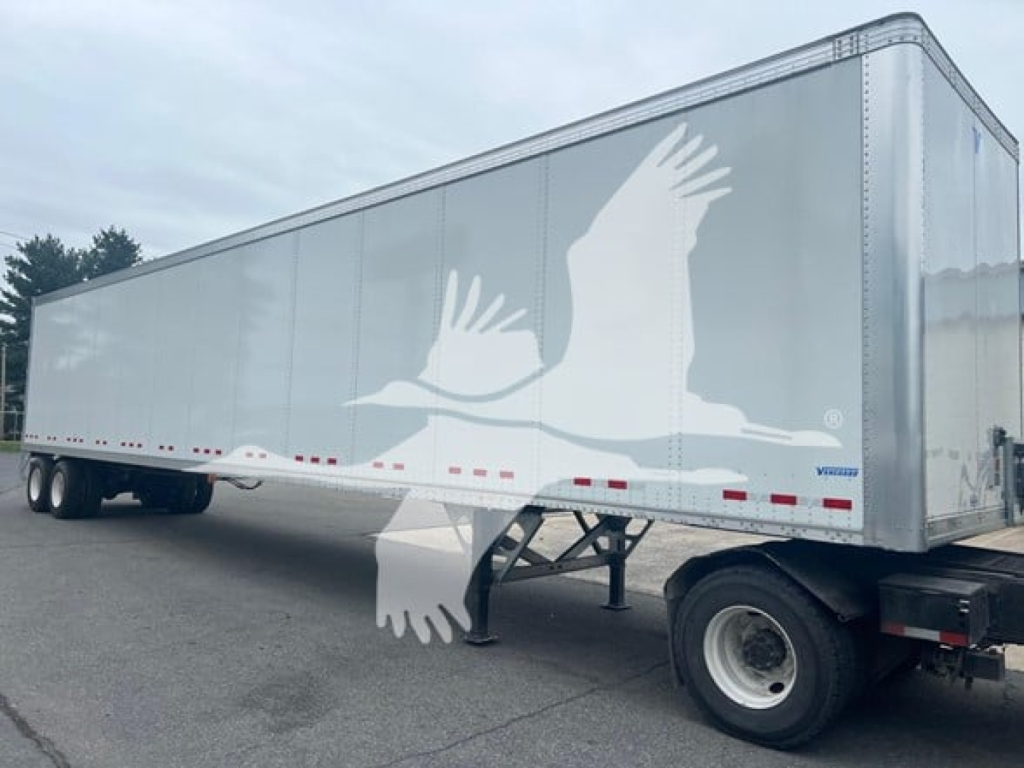 Trailer side view of a Vanguard VXP plate van trailer with a large white bird graphic on silver panels; dual rear axles, hitch, and support legs visible.