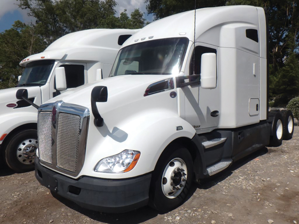 Kenworth T-680 truck in a yard, side view showing aerodynamic sleeper cab, chrome grille, white exterior, dual rear axles, large fuel tanks.