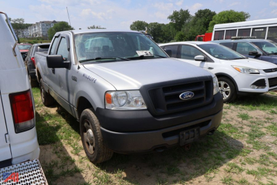 Ford F-150 pickup truck front-left view showing grille with Ford emblem and bumper; silver body with black front bumper and side mirrors; other vehicles in background.