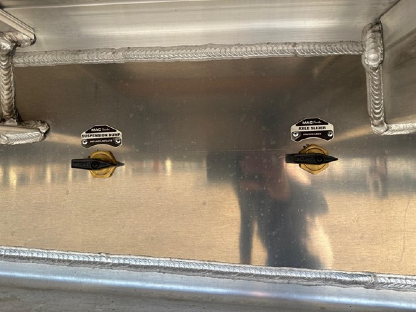 Mac Drop Deck trailer interior showing two valves labeled suspension dump and axle slider, with welded aluminum surface and reflections.