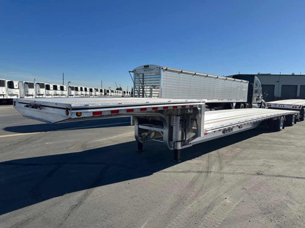 Mac Drop Deck trailer shown in side/ rear angle with flatbed extendable deck and aluminum side rails. visible light bar, wheels, and hitch.