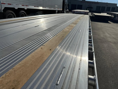 Aluminum drop deck trailer bed with shiny ribbed surface and wood inserts, side rails visible, factory industrial yard in background.