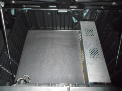 Truck cab interior storage area showing a metal panel with ventilation holes and a large empty floor with a silver power unit on the right.