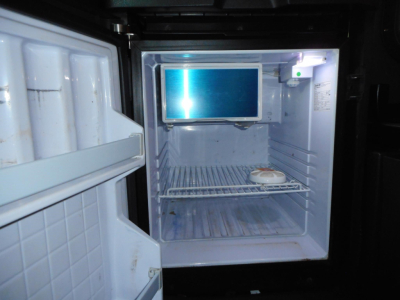 Fridge inside a Freightliner Cascadia sleeper cab showing small monitor mounted on wall and empty freezer shelf area.