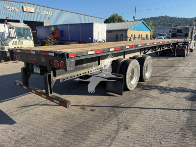 Flatbed trailer viewed from rear-left showing dual rear axles, black frame, red and white reflective tape, Fontaine branding on rear, storage/maintenance yard in background.