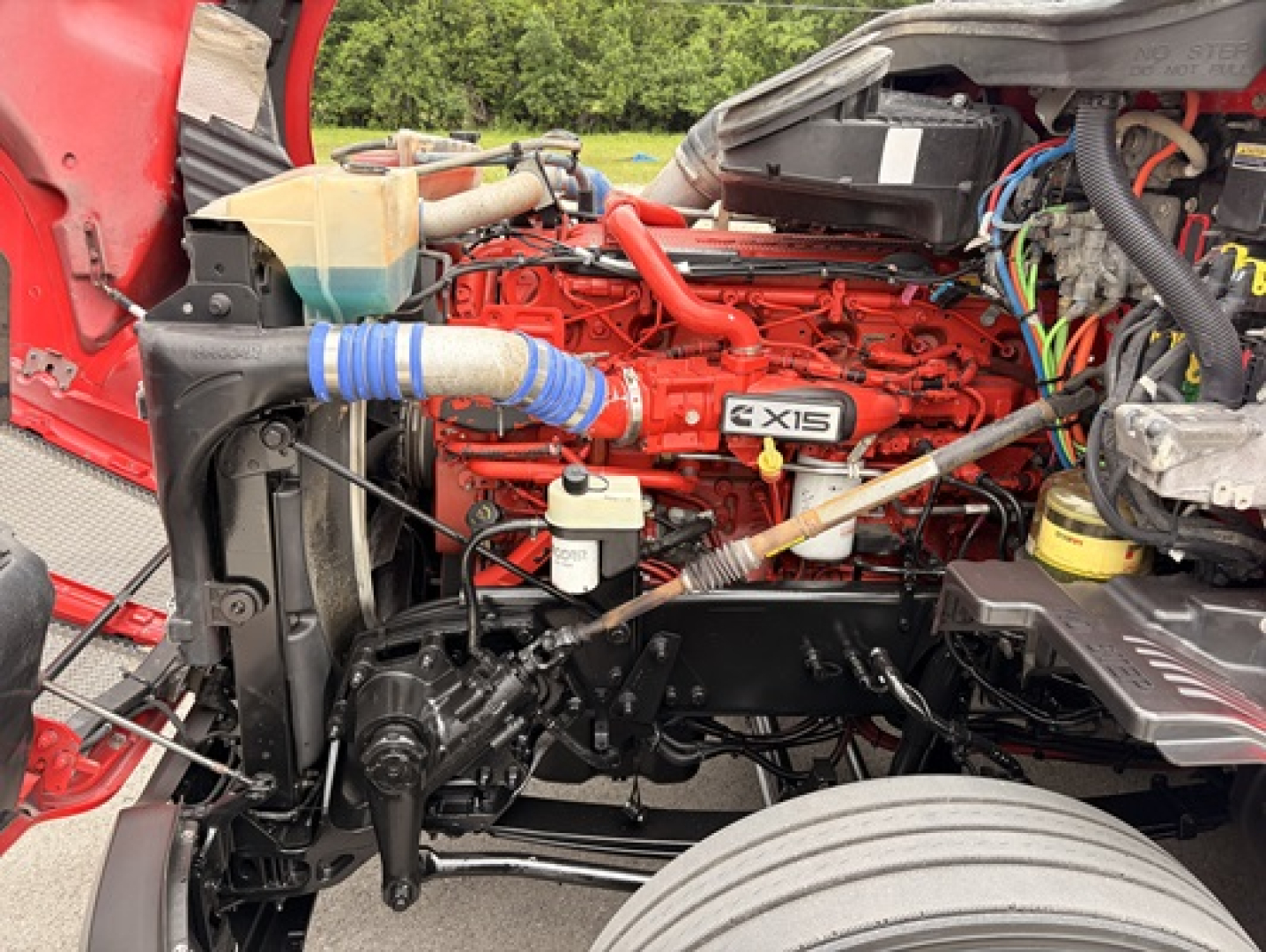Kenworth T680 engine bay showing red Cummins X15 engine with turquoise coolant reservoir, wiring harness, and radiator assembly.