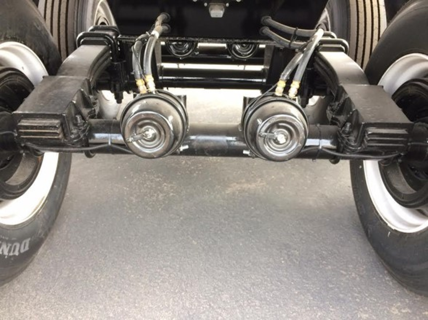 Trailer axle assembly showing dual axles with leaf springs and braking components, tires on both sides, and hydraulic lines in view.