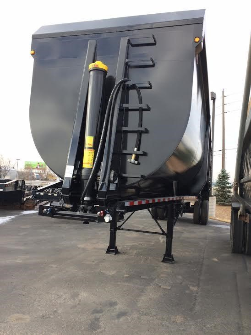 Dump trailer front with hydraulic lift and hitch, black curved dump bed, yellow safety labels, trailer legs deployed.