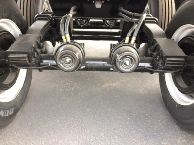 Trailer axle assembly showing dual axles with leaf springs and braking components, tires on both sides, and hydraulic lines in view.